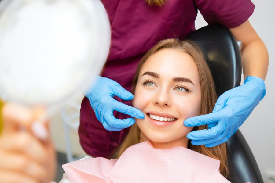 Woman in a dental chair smiling as a gloved provider gently touches her cheeks with a mirror held nearby