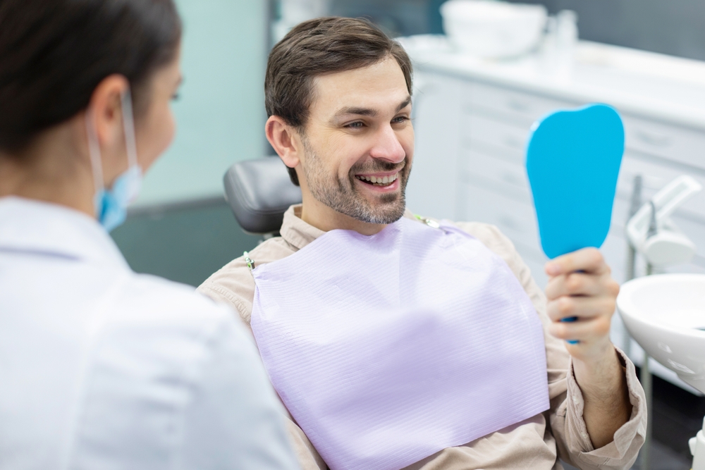 Patient smiling with a hand mirror during a dental visit Smiling patient in a dental bib holding a hand mirror while facing his dentist