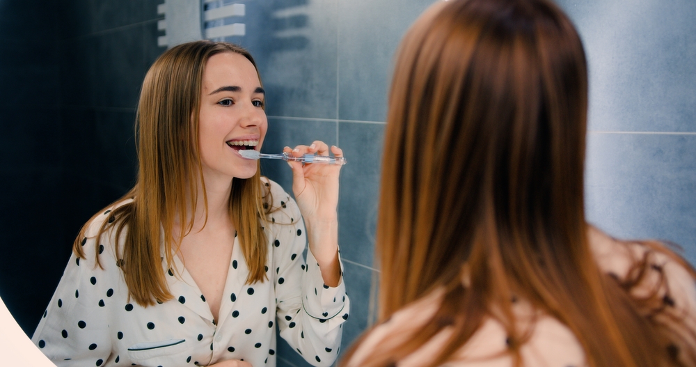 Woman brushing her teeth in front of a bathroom mirror to maintain her smile after teeth whitening