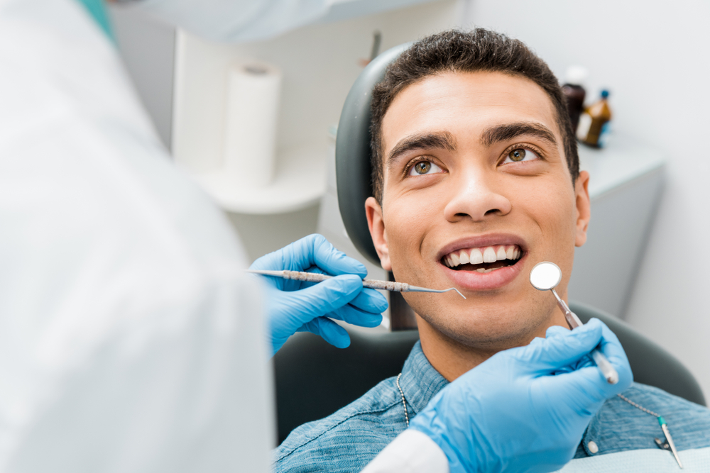 Patient during a dental exam Young man in a dental chair smiling as gloved hands hold an explorer and mirror near his mouth