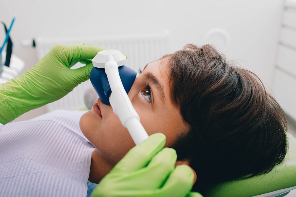Gloved hands placing a nitrous oxide nasal mask on a young patient reclined in a dental chair 