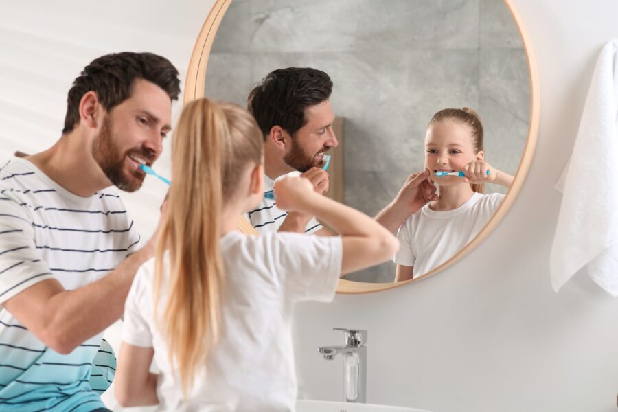 Father and daughter brushing their teeth together in front of a bathroom mirror