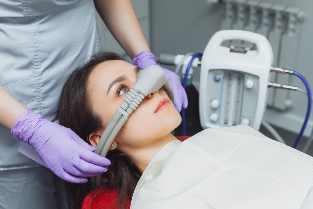 Gloved hands placing a sedation nasal mask on a patient beside a monitoring machine 