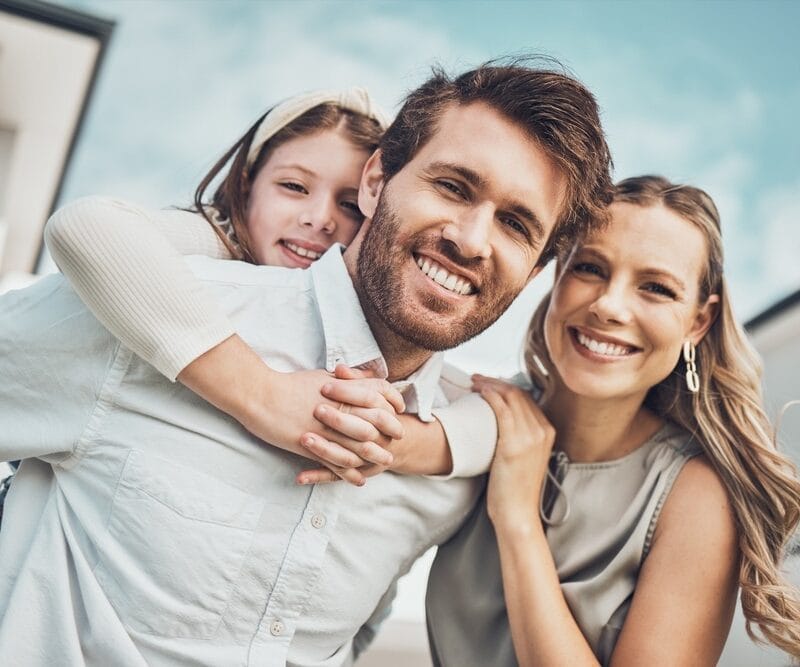 Family of three smiling outdoors with a daughter riding piggyback on her father