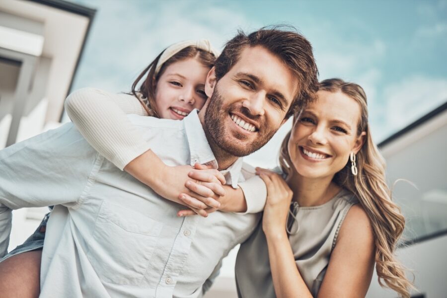Family of three smiling outdoors with a daughter riding piggyback on her father