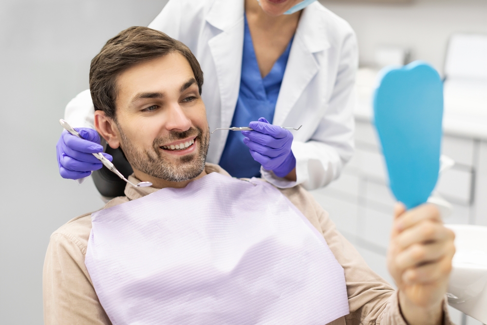 Patient smiling with dentist and hand mirror Smiling patient in a dental bib holding a hand mirror as his dentist holds two instruments behind him