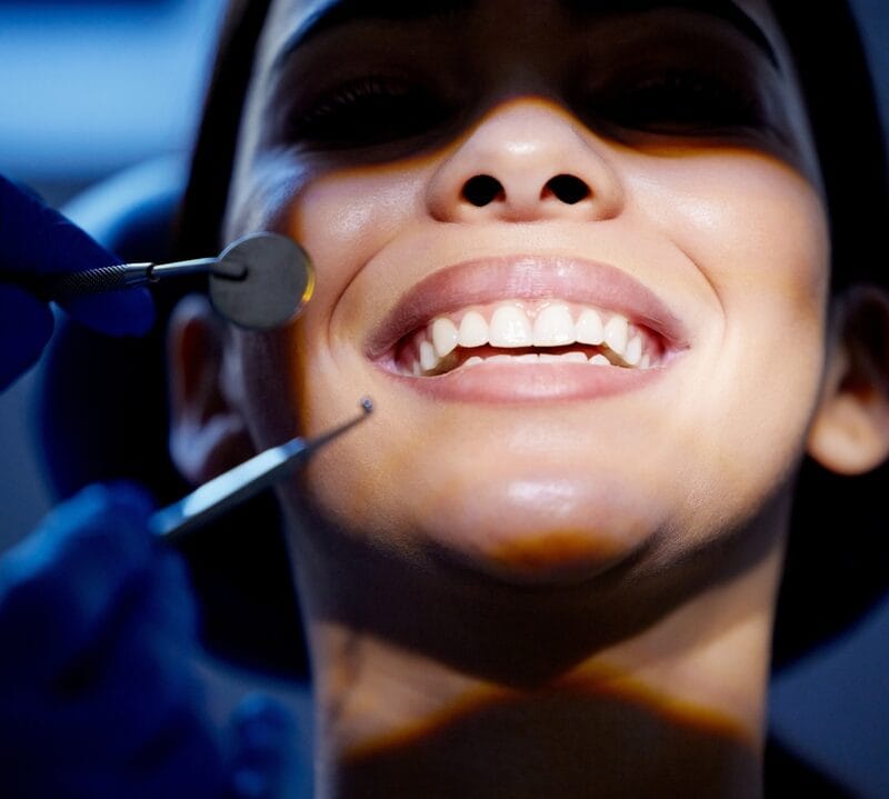 Gloved hands holding a dental mirror and scaler near a patient's smiling teeth
